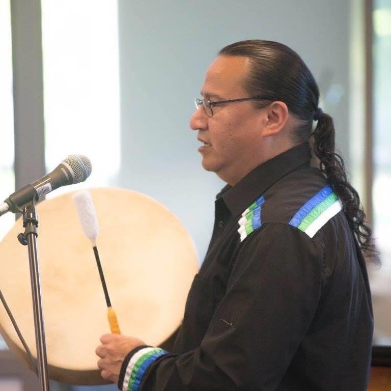 Indigenous man's side profile, playing a hand drum at a microphone.
